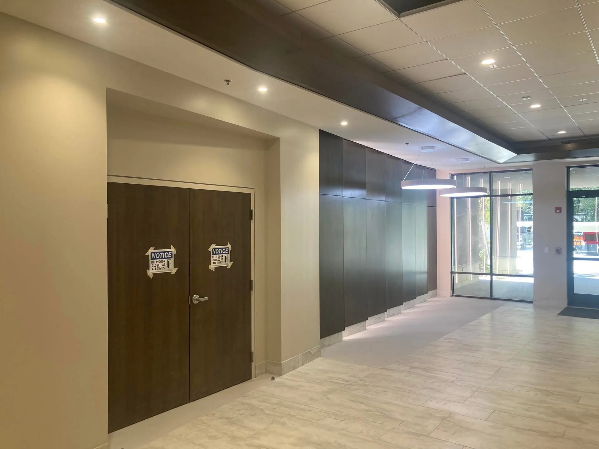 Lobby with dark wood doors, light wall surfaces and a ceiling that steps down toward a glazed corridor.